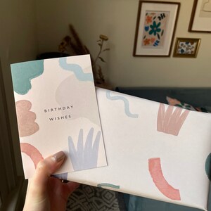 May include: A birthday card and gift box with a pastel abstract design. The card reads "BIRTHDAY WISHES" in black text. The gift box and card feature watercolour-style shapes in shades of blue, pink, and beige. The items are held in a hand.
