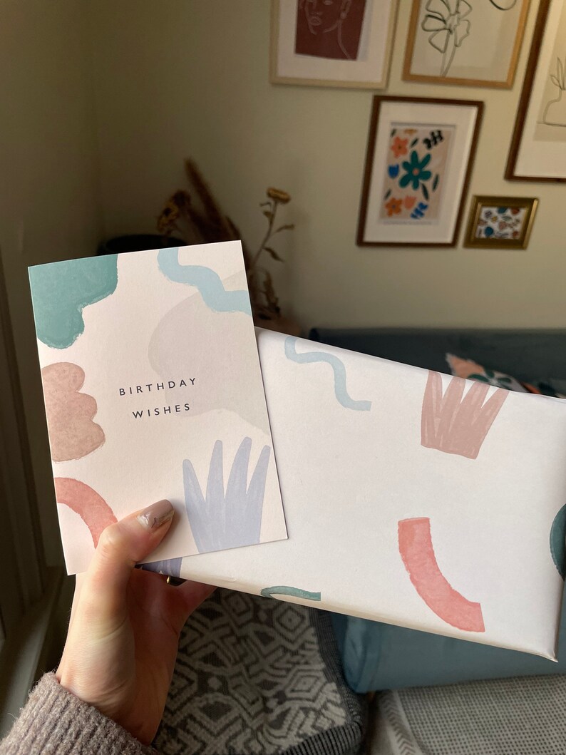 May include: A birthday card and gift box with a pastel abstract design. The card reads "BIRTHDAY WISHES" in black text. The gift box and card feature watercolour-style shapes in shades of blue, pink, and beige. The items are held in a hand.