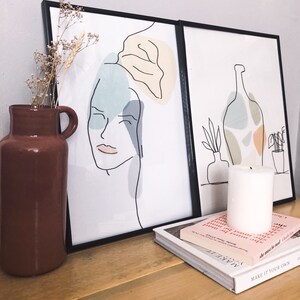May include: Two framed art prints with black frames. One print features a line drawing of a face with abstract shapes in blue, yellow, and grey. The other print shows a bottle and plants. A brown vase with dried flowers, a white candle, and books are on a wooden surface.
