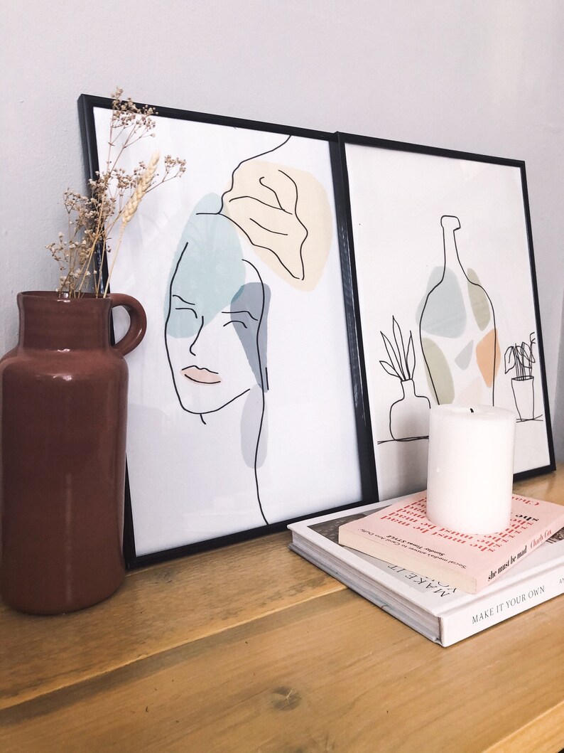 May include: Two framed art prints with black frames. One print features a line drawing of a face with abstract shapes in blue, yellow, and grey. The other print shows a bottle and plants. A brown vase with dried flowers, a white candle, and books are on a wooden surface.