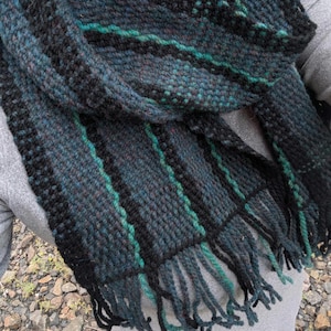 May include: A hand-woven scarf featuring a textured pattern of black, dark teal, and deep blue yarn. The scarf displays vertical stripes in black and teal, with a thick fringe at the bottom edge made of matching black and teal yarn strands.