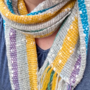 May include: A woven scarf featuring a light gray base with vertical stripes in mustard yellow, bright turquoise, and purple. The fabric has a textured appearance with small white flecks throughout and includes fringed ends in matching colors.