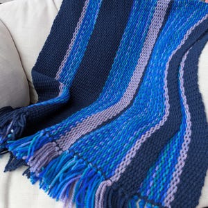 May include: A hand-crocheted throw blanket featuring vertical stripes in shades of navy blue, light blue, and periwinkle. The blanket has a textured stitch pattern and is finished with a fringe along the bottom edge made of matching yarn.
