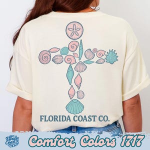 May include: Cream-colored t-shirt featuring a cross design made of seashells in shades of pink and teal. The text "FLORIDA COAST CO." is printed below the cross. The words "Comfort Colors 1717" are printed at the bottom.