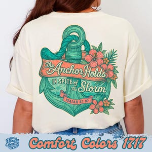 Puede incluir: Camiseta color crema con un diseño de ancla turquesa y el texto "The Anchor Holds In Spite of the Storm" y "Isaías 41:10". El diseño incluye flores de color coral y hojas verdes. La parte inferior de la camiseta tiene el texto "Comfort Colors 1717".
