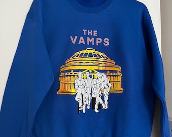Könnte beinhalten: Ein königsblauer Pullover mit einer goldenen und weißen Grafik der Band "The Vamps" vor einem Gebäude. Die Bandmitglieder sind in weißen Silhouetten dargestellt.
