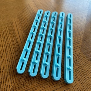 Puede incluir: Seis tiras de plástico azul claro con múltiples agujeros pequeños y un borde redondeado. Las tiras están dispuestas en una fila sobre una superficie de madera.