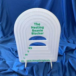 May include: A white plastic beanie blocker with the text "The Nesting Beanie Blocker" in green. Sizes XS, S, M, L, and XL are labeled in green. The brand name "StitchingOriginals" is printed at the bottom.