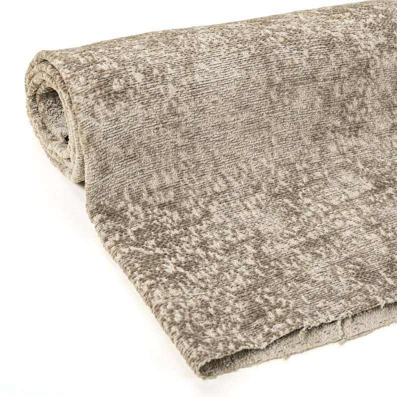 May include: A rolled-up section of textured fabric in shades of beige and brown. The material has a soft, plush appearance with a subtle pattern. The edges of the fabric are visible, showing the layers of the roll. This textile is suitable for upholstery or home decor.