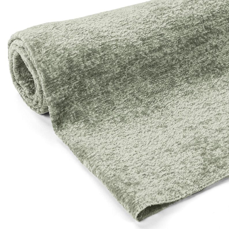 May include: Rolled-up, sage green fabric with a textured, plush surface. The fabric appears soft and has a slight sheen, suggesting a velvet or similar material. The roll reveals the fabric's thickness and construction, suitable for upholstery or decorative use.
