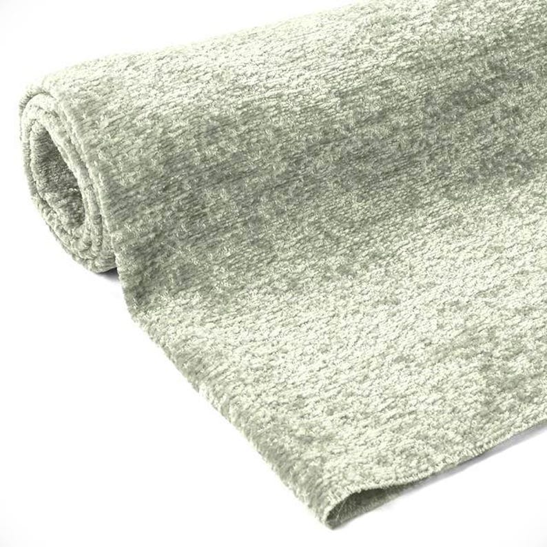 May include: A rolled-up, light green fabric with a textured, plush surface. The fabric has a soft, velvety appearance and a subtle sheen. The material appears to be a heavy weight, suitable for upholstery or drapery.