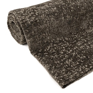 May include: Rolled-up brown area rug with a textured, speckled pattern. The rug has a soft, plush appearance and is made of a thick material. The color is a dark, earthy brown with lighter flecks throughout, creating a subtle visual texture.