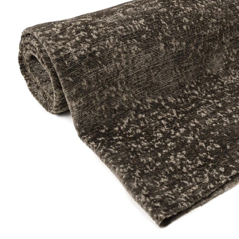 May include: Rolled-up brown area rug with a textured, speckled pattern. The rug has a soft, plush appearance and is made of a thick material. The color is a dark, earthy brown with lighter flecks throughout, creating a subtle visual texture.