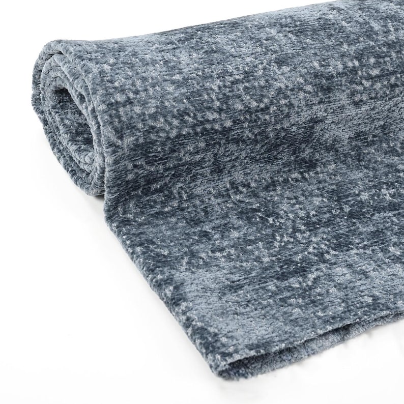 May include: Rolled-up, plush, blue-grey blanket. The soft, textured fabric is visible in a close-up shot. The blanket is rolled up, showing the layers of the material. The color is a muted, cool tone, suitable for home decor.