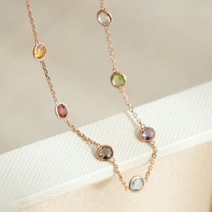 May include: A delicate rose gold necklace featuring a series of colorful gemstones. The gemstones are oval-shaped and set in individual bezels along the chain. The colors include yellow, red, green, purple, brown, and blue. The necklace is displayed on a white surface.