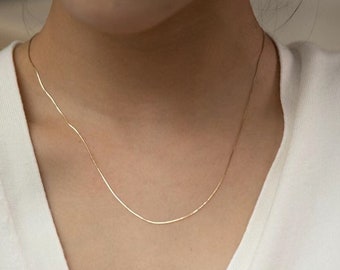 May include: A thin, delicate gold-toned chain necklace worn around the neck. The necklace features a smooth, flexible link design that rests against the collarbone. It is paired with a sleeveless white top that has a deep V-neckline and a single button.
