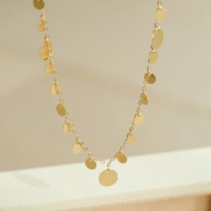 May include: A gold-coloured necklace with a series of small, flat, circular pendants. The pendants are linked by a fine chain, creating a flowing, elegant design. The necklace is displayed against a neutral background.