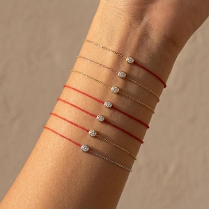 Op de afbeelding: Verschillende delicate armbanden worden op een pols getoond. De armbanden hebben kleine, ronde, heldere edelstenen gezet in goud of zilver. Sommige zijn geregen op rode koorden, andere op fijne gouden kettingen. De achtergrond is neutraal beige.