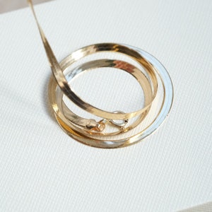 Puede incluir: Un collar de cadena de oro con un dise&ntilde;o de cadena de serpiente. La cadena est&aacute; enrollada sobre una superficie blanca.