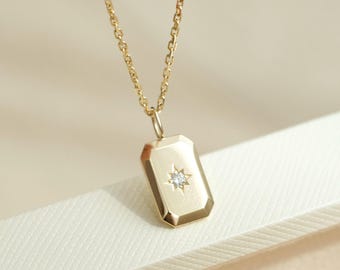 May include: A gold necklace featuring a rectangular pendant with beveled edges. The pendant has a small, clear, round gemstone set in the center, surrounded by an engraved starburst pattern. The pendant hangs from a delicate gold chain link.