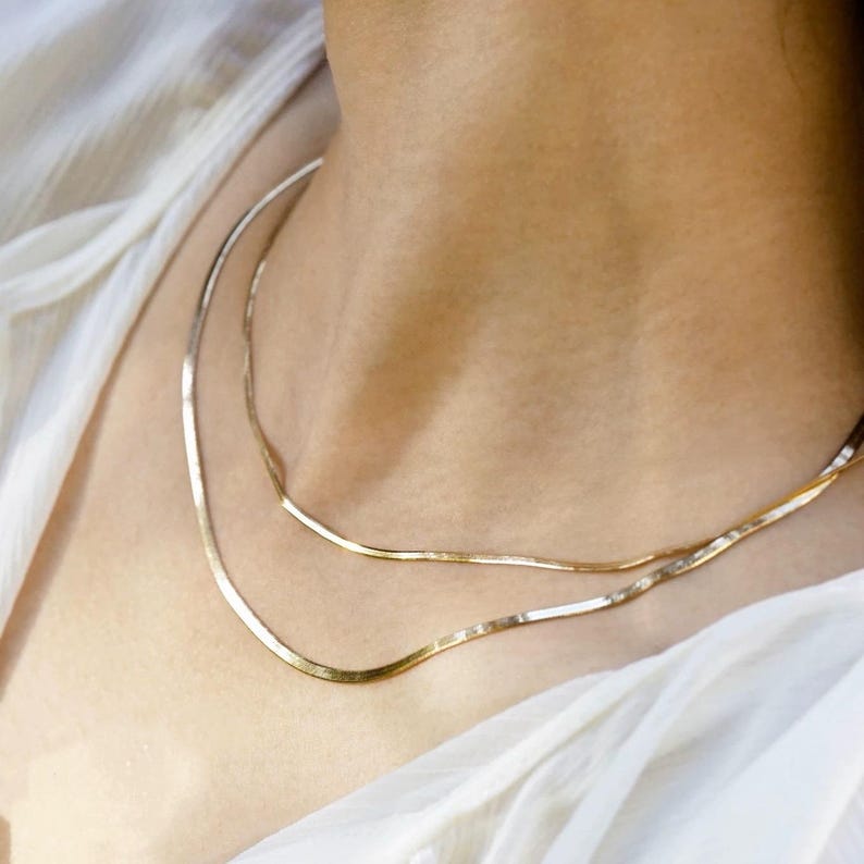 Puede incluir: Dos collares de cadena de oro superpuestos en el cuello de una persona. Las cadenas son finas y tienen un acabado delicado y brillante.