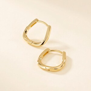 May include: Two gold-colored hoop earrings with a unique, slightly twisted design. The earrings have a polished, reflective surface, and a simple clasp closure. The earrings are displayed on a light beige background.