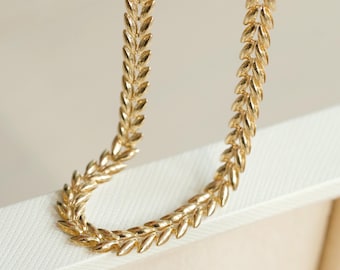 May include: Close-up of a gold-colored necklace with a leaf design. The necklace is made of small, detailed leaves arranged in a repeating pattern. The necklace is resting on a white surface.