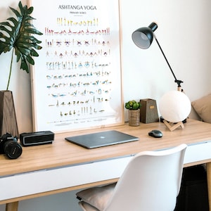 Puede incluir: Un escritorio blanco con una tapa de madera y una silla blanca. Hay una computadora port&aacute;til, una l&aacute;mpara, una l&aacute;mpara de luna, una planta y un p&oacute;ster enmarcado que dice "Ashtanga Yoga Primary Series" en el escritorio.
