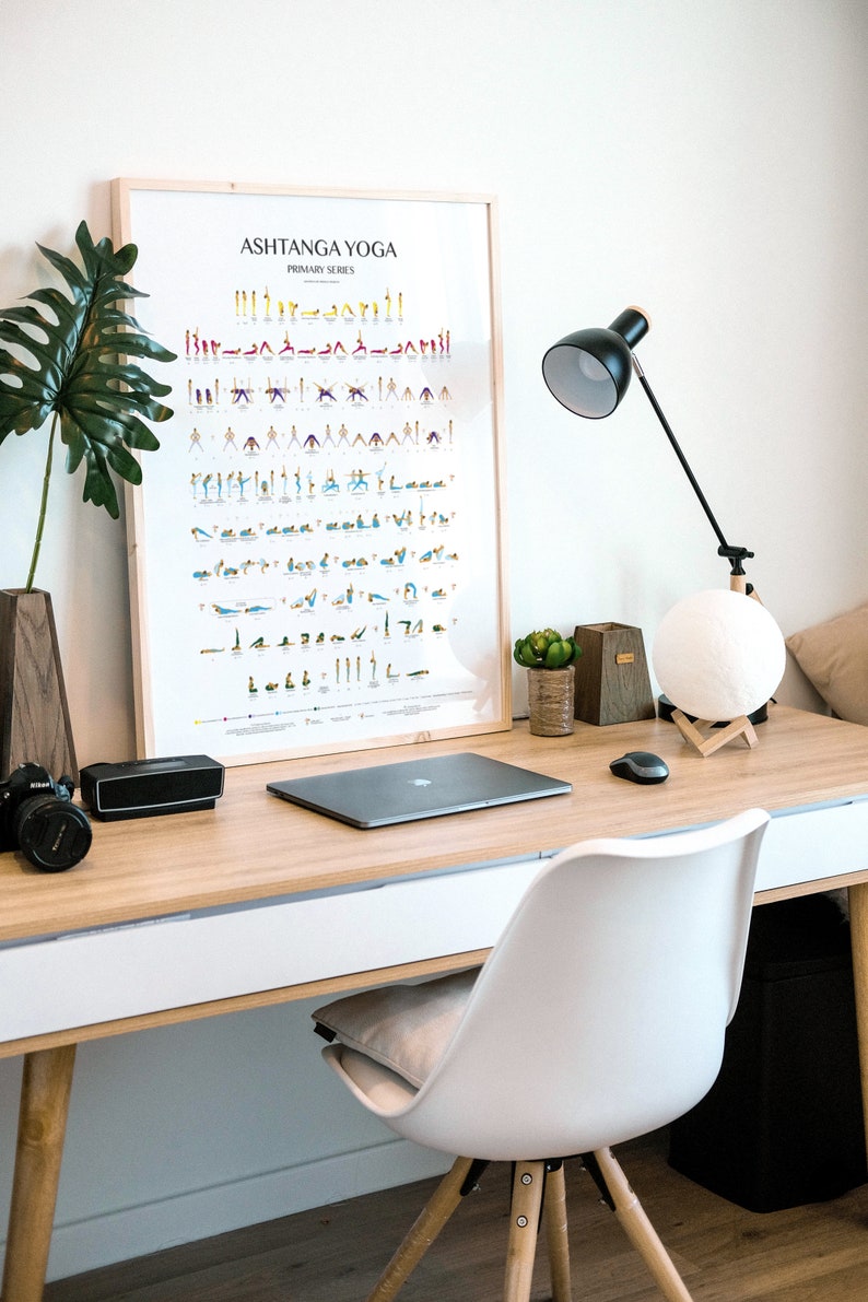 Puede incluir: Un escritorio blanco con una tapa de madera y una silla blanca. Hay una computadora port&aacute;til, una l&aacute;mpara, una l&aacute;mpara de luna, una planta y un p&oacute;ster enmarcado que dice "Ashtanga Yoga Primary Series" en el escritorio.