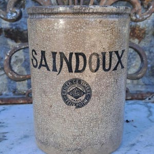 Op de afbeelding: Een cilindervormige beige keramische pot met een opvallende craquelé glazuurafwerking. Het woord SAINDOUX staat in zwarte letters op de voorzijde gedrukt, boven een ronde zwarte stempel met de tekst EXIGER CE TIMBRE FELIX POTIN PARIS.