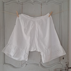 May include: White vintage bloomers hanging on a clothesline. The bloomers have a ruffled hem with delicate cut-out detailing. The background is a light blue door with a rustic, aged appearance.