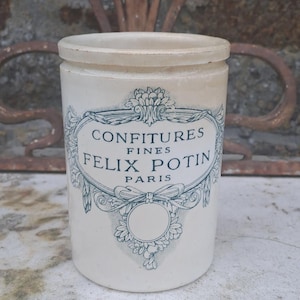 May include: A cream-coloured ceramic jar with a lid, bearing the text "CONFITURES FINES FELIX POTIN PARIS" in blue. The jar is on a stone surface. The jar is a vintage item.
