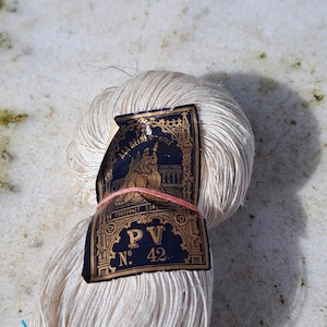 Op de afbeelding: Een bolletje crèmekleurig garen met een vintage label. Het label is donkerblauw en goudkleurig met de tekst "A la Reine" en "PV No. 42". Het garen is gebonden met een roze draad.
