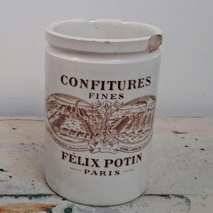 Peut inclure: Un pot en céramique blanc avec des inscriptions et des illustrations marron. Le pot porte les inscriptions "CONFITURES FINES" et "FELIX POTIN PARIS". Les illustrations représentent des bâtiments et des arbres. Le bord du pot est légèrement ébréché.