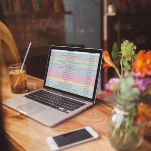 May include: A laptop computer with a bright, colorful spreadsheet open on the screen. The spreadsheet lists various assignments and due dates. The laptop is on a wooden table with a glass of iced tea and a smartphone.