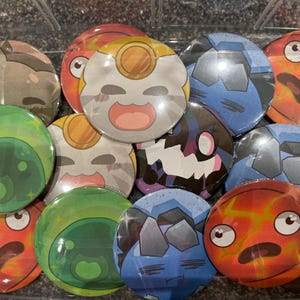 May include: A collection of 11 round, colourful buttons featuring various cartoon characters and designs. The buttons are made of metal and have a shiny finish. The characters include a slime, a ghost, a cat, and a red creature with a lightning bolt.