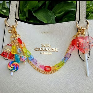May include: Cream handbag with gold-tone hardware and a rainbow charm. The charm includes colourful beads, a lollipop, and a heart-shaped lollipop. The bag features a gold Coach logo.