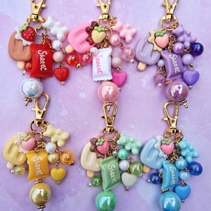 May include: Six colorful keychains with gold-tone clasps. Each keychain features a popsicle, candy, and heart charms. The keychains have a variety of colors, including brown, pink, purple, yellow, green, and blue. Each keychain has a charm that says "Sweet".