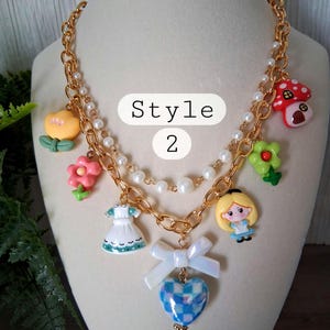 Pu&ograve; includere: Collana a catena color oro con ciondoli multipli, tra cui un fungo, un fiore, un vestito, un cuore e un personaggio di Alice nel Paese delle Meraviglie. La collana ha una fila di perle e il testo "Style 2".