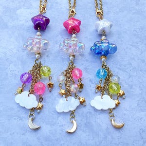 May include: Three gold chain necklaces with star, cloud, and moon charms. Each necklace features a different colour scheme: purple, pink, and blue. The charms are made of iridescent plastic and have a whimsical, celestial theme.