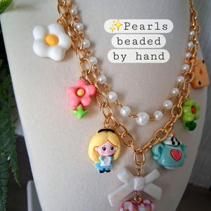 Pu&ograve; includere: Una collana dorata con perle finte bianche e ciondoli colorati. I ciondoli includono fiori, un personaggio dei cartoni animati, una teiera e un ciondolo a forma di cuore. Il testo "Pearls beaded by hand" &egrave; visibile.