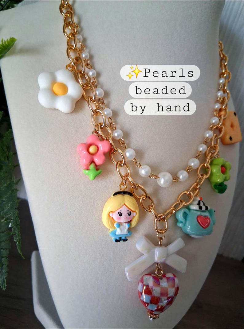 Pu&ograve; includere: Una collana dorata con perle finte bianche e ciondoli colorati. I ciondoli includono fiori, un personaggio dei cartoni animati, una teiera e un ciondolo a forma di cuore. Il testo "Pearls beaded by hand" &egrave; visibile.