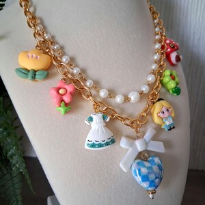 Pu&ograve; includere: Una collana dorata con diversi ciondoli. I ciondoli includono un fiore giallo, un fiore rosa, un fungo rosso, una rana verde, un vestito bianco, un personaggio di Alice nel Paese delle Meraviglie e un cuore a scacchi blu e bianco. La collana ha anche una fila di perle bianche.