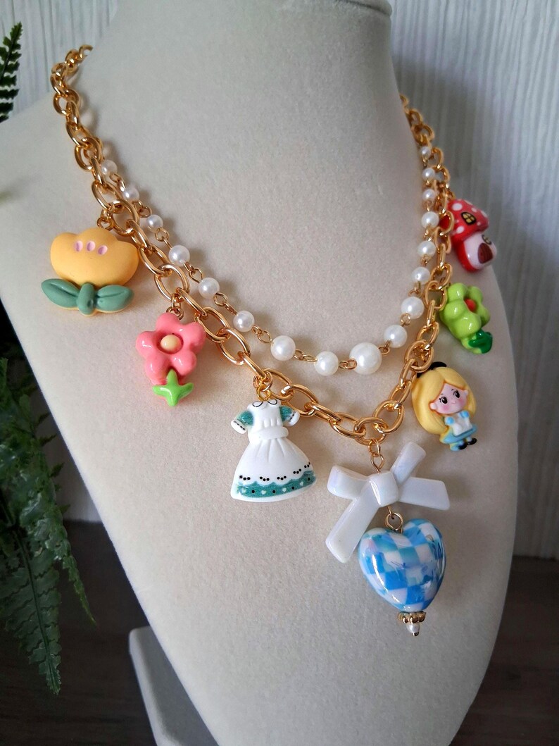 Pu&ograve; includere: Una collana dorata con diversi ciondoli. I ciondoli includono un fiore giallo, un fiore rosa, un fungo rosso, una rana verde, un vestito bianco, un personaggio di Alice nel Paese delle Meraviglie e un cuore a scacchi blu e bianco. La collana ha anche una fila di perle bianche.
