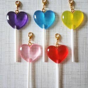 May include: Five heart-shaped lollipops in shades of purple, blue, yellow, pink, and red. Each lollipop has a gold-colored clasp and a white stick. The lollipops are arranged against a light gray background.