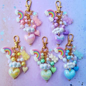 May include: Five keychains with gold-colored clasps, each adorned with rainbow, cloud, and heart charms. The charms feature pastel colors like pink, yellow, and blue, with iridescent heart pendants. The keychains are arranged on a gradient background.