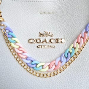 Puede incluir: Bolso de mano de cuero blanco con herrajes dorados y una correa de cadena arcoíris pastel. El bolso presenta el logotipo de Coach en letras doradas. La correa de cadena está hecha de eslabones entrelazados en tonos rosa, amarillo, azul y morado.