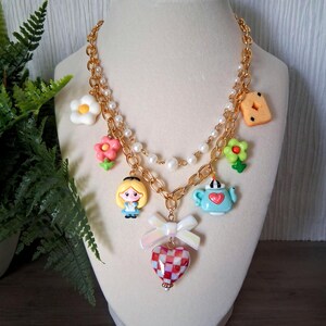 Pu&ograve; includere: Una collana a catena color oro a pi&ugrave; fili con una fila di perle bianche finte e vari ciondoli tra cui un personaggio biondo, una teiera azzurra, un fiocco bianco, un cuore a scacchi rossi e bianchi e fiori colorati.