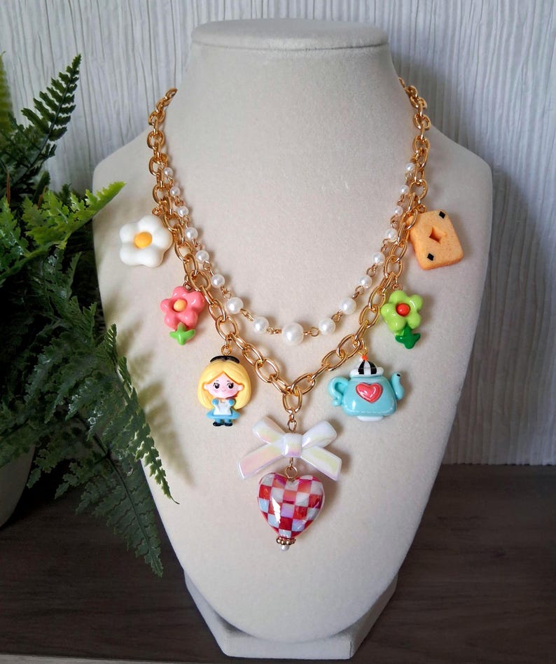 Pu&ograve; includere: Una collana a catena color oro a pi&ugrave; fili con una fila di perle bianche finte e vari ciondoli tra cui un personaggio biondo, una teiera azzurra, un fiocco bianco, un cuore a scacchi rossi e bianchi e fiori colorati.