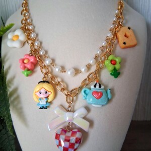 Pu&ograve; includere: Una collana color oro con diversi ciondoli, tra cui un personaggio dei cartoni animati, fiori, una teiera e un biscotto. La collana ha una fila di perle e un ciondolo a forma di cuore con un motivo a scacchi rosso e bianco. Un fiocco bianco &egrave; attaccato al cuore.